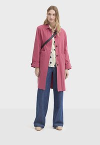 Manteau long rose avec col, poches devant et boutons, porté sur une chemise blanche à pois noirs et associé à un jean bleu à jambes larges.
