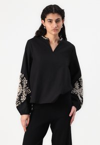 Blusa nera con scollatura a fessura, caratterizzata da maniche lunghe con ricami floreali beige e polsini arricciati. Tessuto morbido con una vestibilità rilassata.