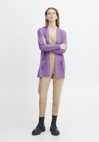 Fialový pletený cardigan s kapsami, vrstvený přes béžový top, kombinovaný s béžovými kalhotami a černými šněrovacími botami. Hladká textura.