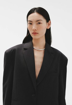 Blazer negro de corte oversized con solapa de muesca, cierre de un botón y bolsillo confeccionado, llevado sobre una blusa de escote en V profundo; textura suave.