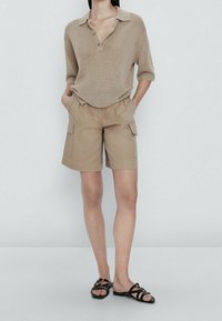 Beige knitted polo top with a loose fit and a three-button placket, paired with light beige cargo shorts featuring side pockets. Black sandals.