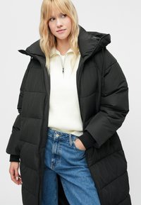 Fekete, kapucnis puffermellény, steppelt textúrával és teljes cipzárral. Fehér bordázott pulóver és kék farmer felett viselve.