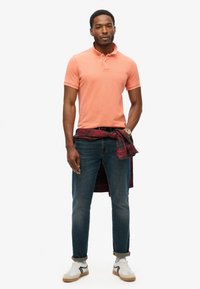 Heren polo met korte mouwen in perzik, gecombineerd met donkere jeans. Een rood geruit overhemd om de taille gebonden, en witte sneakers met grijze accenten.