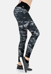Hoch geschnittene schwarze Leggings mit einem strukturierten weißen abstrakten Muster, hergestellt aus dehnbarem Stoff, kombiniert mit dunklen Sport Schuhen.