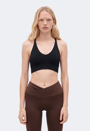 Un mannequin blonde se tient face à l’objectif, vêtue d’une brassière de sport noire et d’un legging marron foncé, sur un fond clair uni.