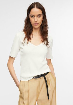 Woman wearing white short-sleeve top with scalloped neckline and beige high-waist pants with a black braided belt, hands in pockets.