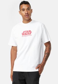 Bílý bavlněný tričko s červenou grafikou "Star Wars: Impérium vrací úder" na přední straně. Krátké rukávy a kulatý výstřih.