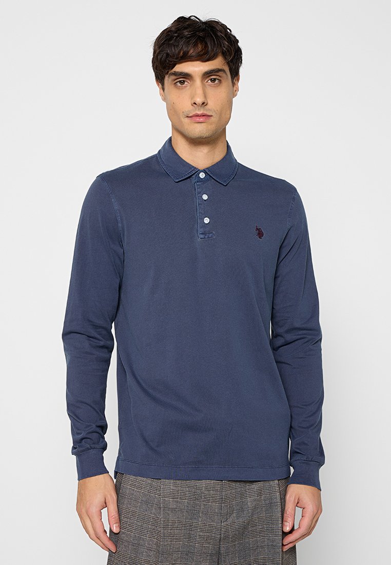 Mann in einem navyblauen Langarm-Polohemd mit drei Knöpfen und grauen karierten Hosen, steht vor einem schlichten Hintergrund.