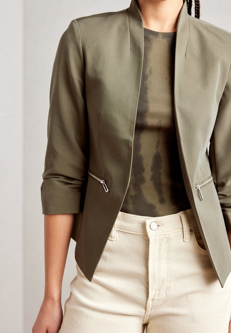 Oliwkowa marynarka z szerokim kołnierzem, długimi rękawami i zamykanymi na zamek kieszeniami, połączona z prążkowanym, ciemnooliwkowym topem i jasnobeżowymi spodniami.