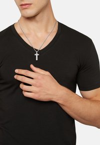 Sort v-hals t-shirt med en tætsiddende design; model iført en sølvkæde med et korsvedhæng. Hånden placeret på brystet.