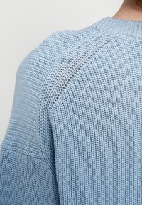 Pull tricoté bleu clair avec un motif texturé, un col arrondi et un détail en maille ajourée le long des coutures d'épaule.