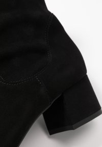 Stivaletto in suede nero con tacco a blocco e cuciture visibili su uno sfondo bianco.