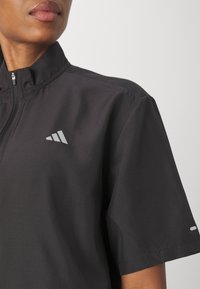 T-shirt sportif noir à demi-fermeture éclair avec manches courtes, arborant un logo réfléchissant et un tissu lisse et léger. Design simple et fonctionnel.