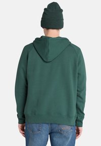 Felpa verde con cappuccio e tasca a marsupio, polsini e orlo a coste, abbinata a un berretto di lana verde coordinato. Jeans in denim visibili.