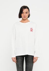 Λευκό πουκάμισο με μακριά μανίκια και στρογγυλή λαιμόκοψη, με κόκκινο γραφικό "NY Coffee" στο αριστερό στήθος. Φορεμένο με σκούρο τζιν παντελόνι.