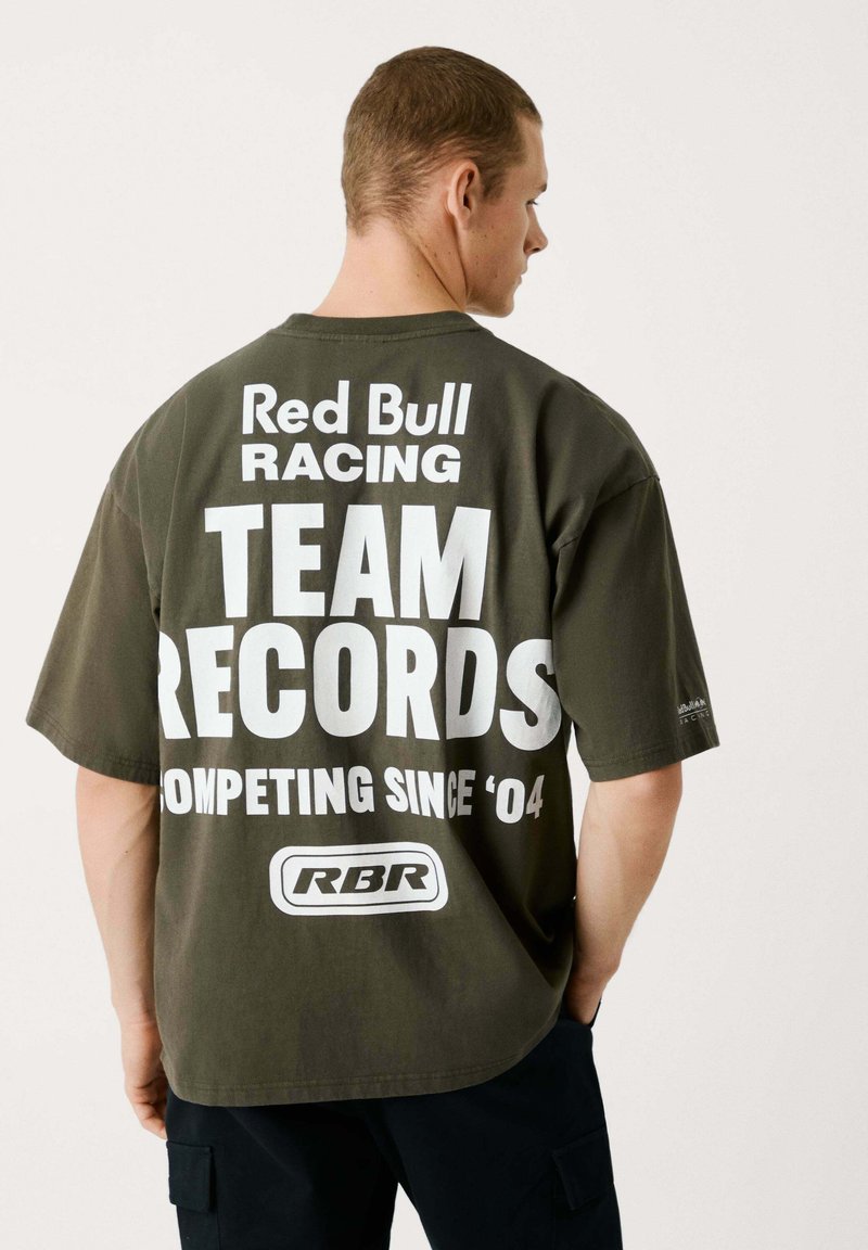 Olivza zaļā oversized T-krekls, izgatavots no kokvilnas, ar izteiksmīgiem baltajiem uzrakstiem aizmugurē: "Red Bull Racing KOMANDA REKORDI SACENŠOTS NO '04."