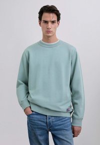 Sudadera verde menta con cuello redondo, puños y dobladillo acanalados. Presenta una pequeña etiqueta de tela en la parte inferior izquierda. Combinada con jeans azules.
