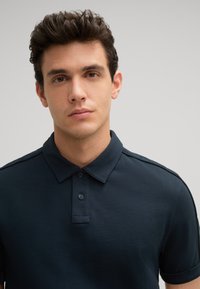 Dunkelblaues Poloshirt mit Kragen, drei Knöpfen und kurzen Ärmeln. Aus glattem Stoff gefertigt, mit taillierter Passform und dezenten Details an den Schultern.