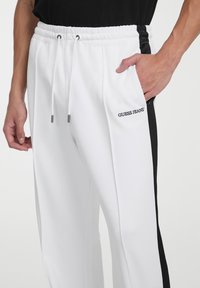 Weiße Jogginghose mit schwarzem Seitenstreifen, elastischem Bund, Kordelzugverschluss und gesticktem "GUESS JEANS"-Logo auf der Vorderseite.