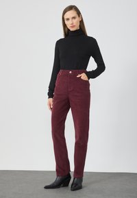 Hoge taille bordeaux corduroy broek met voorkruiszakken en een metalen knoop, gecombineerd met een aansluitende zwarte colsjaal en zwarte haklaarzen.