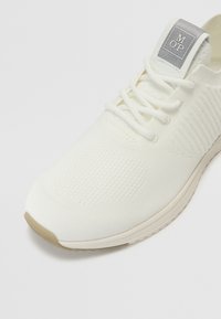 White athletic shoe with a textured knit upper, flat laces, and a soft rubber outsole. Features a branded tongue tag with gray accents.