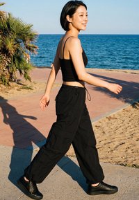 Young woman wearing black crop top and cargo pants walking on a beachside path near palm trees with ocean in the background on a sunny day.