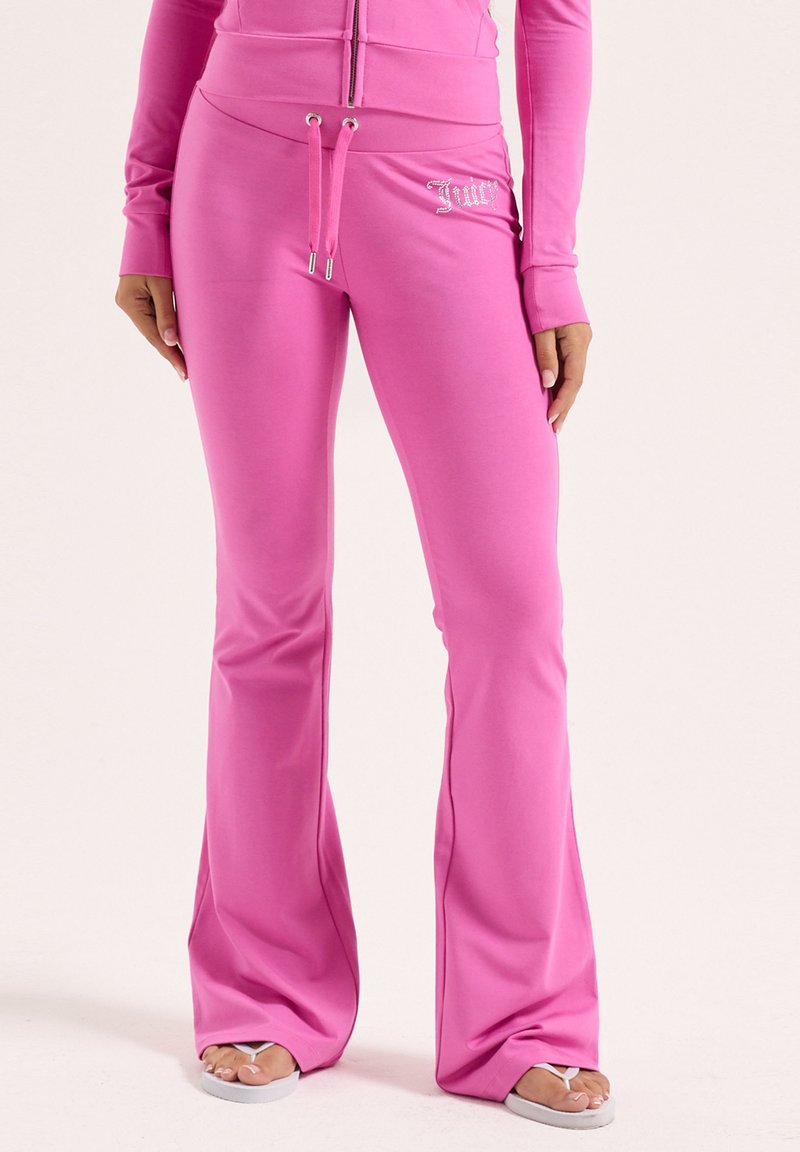 Femme portant un pantalon évasé rose vif avec une taille à cordon et un logo "Juicy" en strass, associé à des tongs blanches.