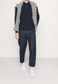 Polo navy combinado con pantalones de mezclilla azul oscuro y zapatillas blancas. Suéter a rayas casualmente colocado sobre los hombros, hecho de tela ligera.