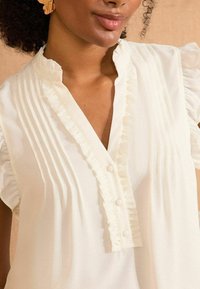 Vit blus med veckad tyg och volangdetaljer längs kragen och knappslån, med korta, puffiga ärmar.
