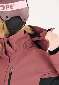 Rosa skidjacka med hög krage och dragkedjefickor, bärs med en svart hjälm. Materialet verkar vara vattenavvisande med en slät textur.