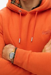 Persona vistiendo una sudadera con capucha naranja de Superdry con puños de camisa a cuadros visibles, brazos cruzados y un reloj de pulsera con correa de cuero marrón en la muñeca izquierda.