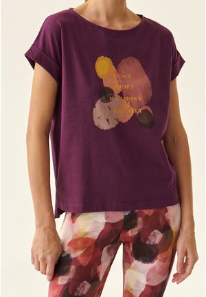 Vrouw draagt een paarse blouse met abstracte oranje en gele vormen en de woorden vrede, rust, harmonie, sereniteit in gouden letters.