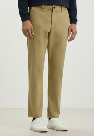 Chino khaki realizzati in tessuto morbido, con un design a gamba dritta, tasche frontali e chiusura a un solo bottone, abbinati a scarpe da ginnastica bianche.