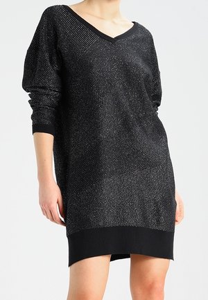 Femme portant une robe en maille noire à manches longues avec un léger scintillement et un décolleté en V, arrivant à mi-cuisse.