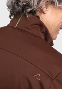 Braune Jacke mit hohem Kragen, grünen Zickzacknähten und einem kleinen gestickten Logo auf der Rückseite in der Nähe des Nackenbereichs.