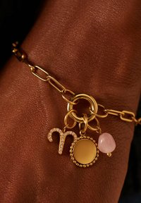Gouden kettingarmband met een ronde hanger, een rozenkwartscharme en een sprankelend sterrebeeldsymbool, tegen een gestructureerde huidachtergrond.