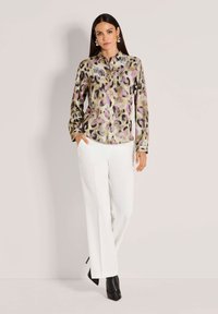 Woman wearing a leopard-print long-sleeve blouse with purple accents, white pants, black ankle boots, and gold hoop earrings, standing.