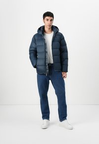 Dark navy puffer jacket with a hood, featuring a quilted design and front zipper. Paired with light gray t-shirt and blue jeans, white sneakers.