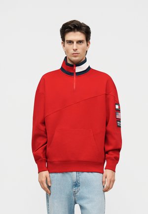 Młody mężczyzna w czerwonej bluzie z zamkiem do ćwiartki i naszywkami na lewym rękawie oraz jasnoniebieskich dżinsach, stojący na tle jednolitego tła.