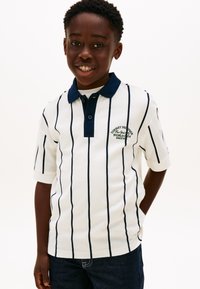 Λευκό πόλο πουκάμισο με κάθετες ναυτικές ρίγες, γιακά με κουμπιά και κεντημένο λογότυπο. Κοντά μανίκια και υφασμάτινη υφή. Φοριέται με μπλε τζιν.