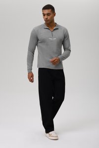 Sudadera gris con cremallera y mangas largas, con un logotipo blanco en el pecho, combinada con pantalones negros de corte holgado y zapatillas beige.