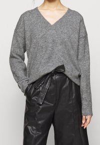 Maglione grigio in lana a V con spalle scese, abbinato a pantaloni in pelle nera a vita alta con dettaglio della cintura legata.