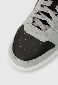 Pilka ir juoda sportinė avalynė su tekstūruota odine viršutine dalimi, perforuotu pirštų apdangalu, plokščiais pilkais raišteliais ir balta guminė pado dalimi.