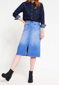 Jeanskjol med medellängd, med en ombre-effekt från mörkblå till ljusblå, och en framficka. Kombinerad med en mörk skjorta med knappar.