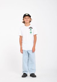T-shirt en coton blanc avec un design graphique sur la poche gauche de la poitrine, associé à un jean bleu clair ample et à des baskets noires. Casquette noire avec un design.