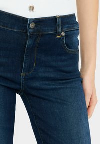 Jeans de mezclilla azul oscuro con un corte clásico, costuras visibles y acentos de hardware metálico. Textura suave con ligeros detalles de desgaste.