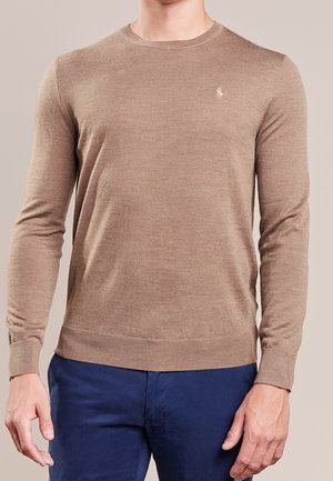 Homme portant un pull à col rond marron clair à manches longues avec un petit logo sur la poitrine et un pantalon bleu foncé.