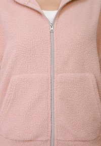 Hanorac roz deschis din fleece cu fermoar, suprafață texturată, două buzunare frontale și un fermoar argintiu. Are un design cu guler înalt.