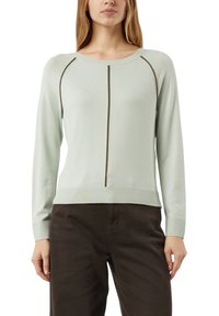 Kobieta ubrana w jasnozielony sweter z długim rękawem z ciemnymi pionowymi szwami, w połączeniu z ciemnobrązowymi spodniami.