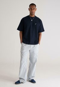 Navy short-sleeve t-shirt with white logo, paired with wide, striped blue and white pants. Model wears navy sneakers.